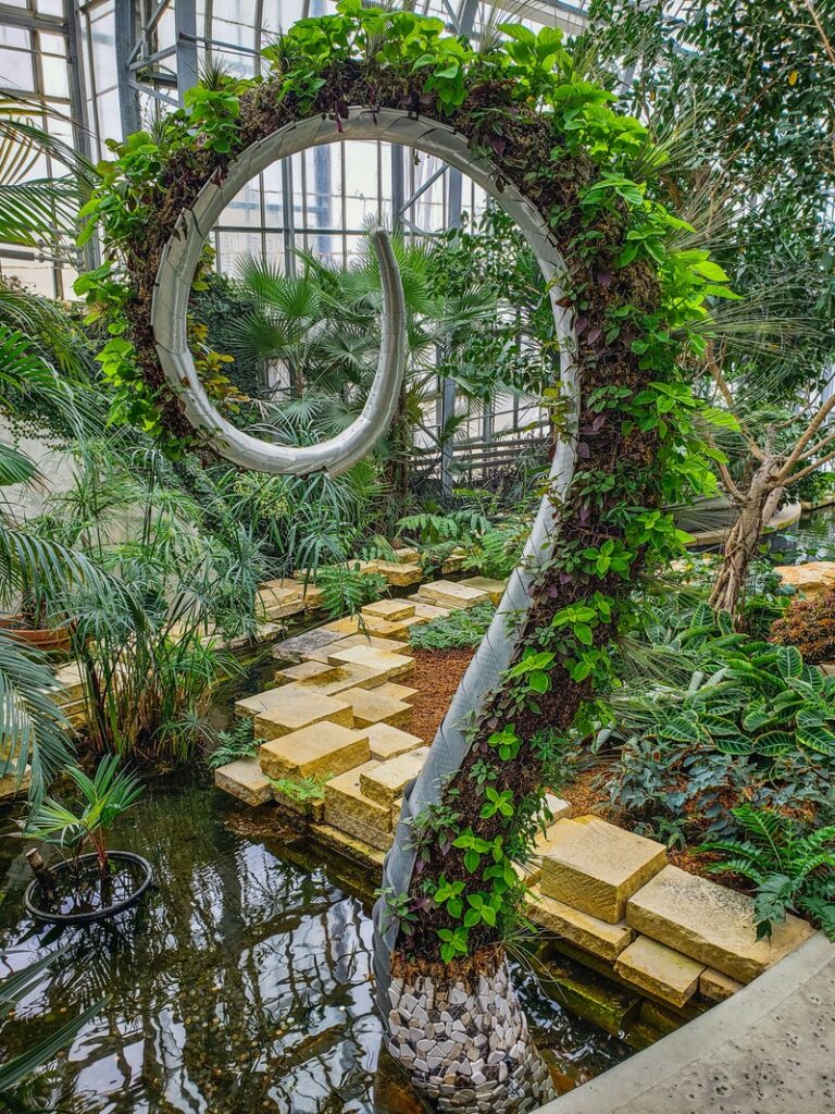 Une grande sculpture en métal en forme de point d'interrogation, recouverte de plantes vertes, se dresse à côté d'un étang dans une serre remplie de végétation tropicale et de chemins en pierre.