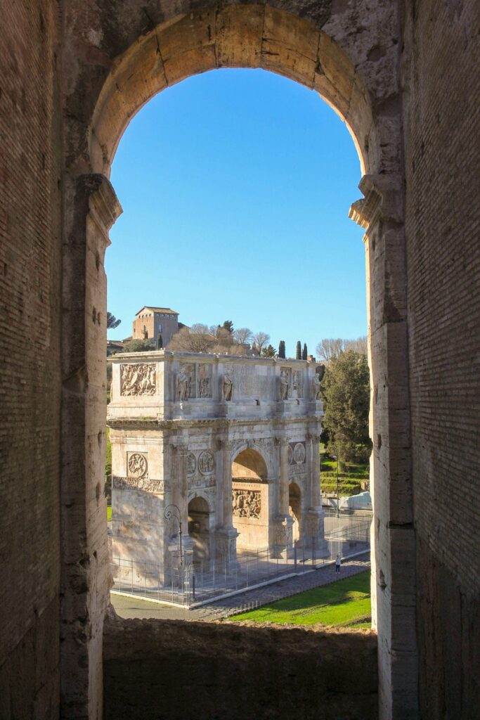 Vue à travers une arche en pierre encadrant l'Arc de Constantin, un ancien arc de triomphe romain, avec des arbres et un ciel bleu clair en arrière-plan.
