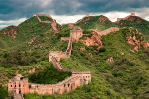 La Grande Muraille de Chine s'étend sur un terrain verdoyant et vallonné sous un ciel nuageux. Des tours de guet en pierre sont visibles tout au long du mur.