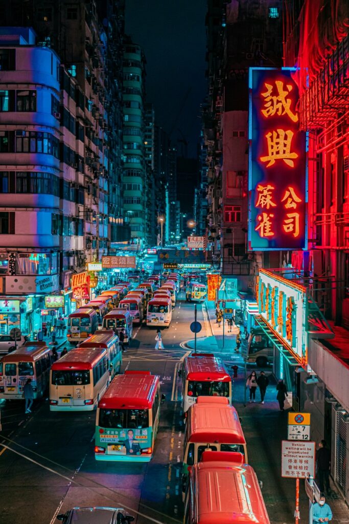 Des enseignes au néon colorées illuminent une rue urbaine animée la nuit, avec de nombreux bus, voitures et piétons se déplaçant entre de grands immeubles.